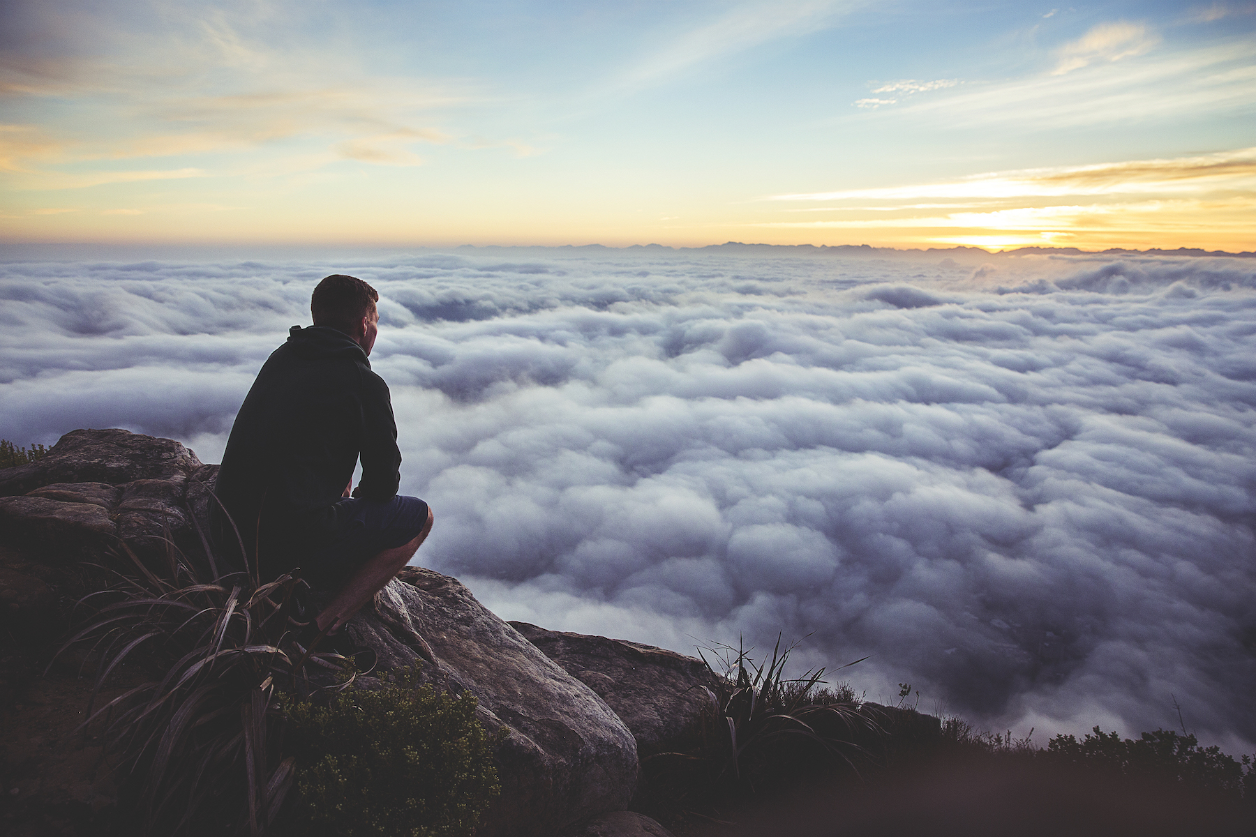 Man on a mountain cliff, looking onward over the clouds below. 